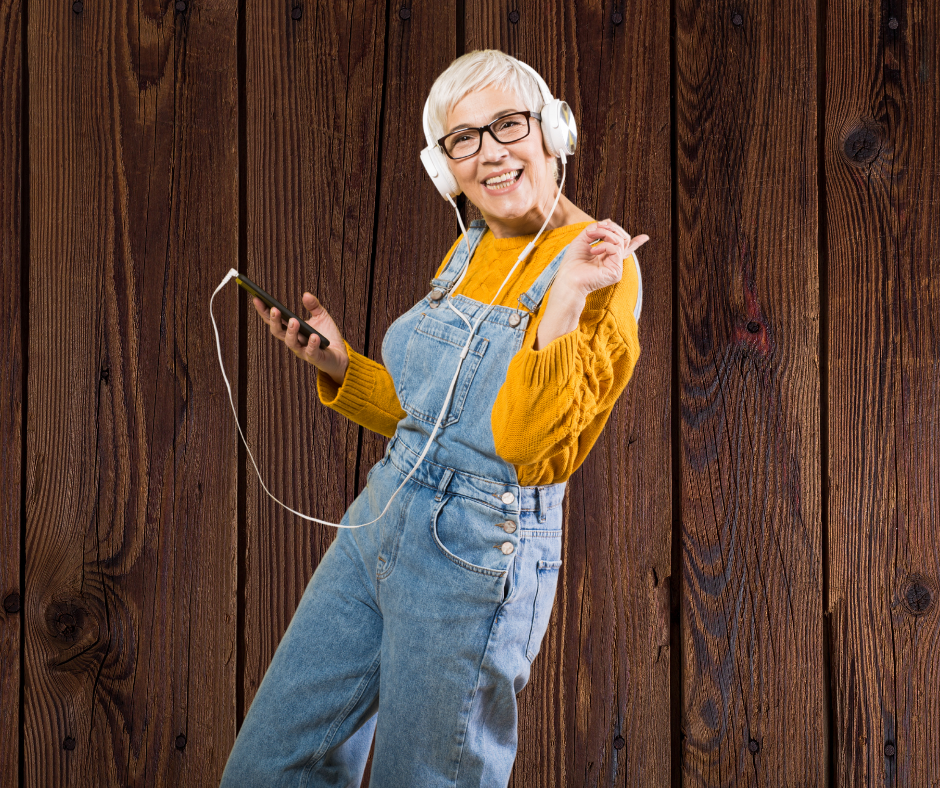eine Frau mit weißen Haaren und Brille trägt einen Ockerfarbenen Pulli und jeanslatzhose, Kopfhörer angeschlossen am Handy, sieht aus als würde sie tanzen auch so geht #zeigdichbunt heute