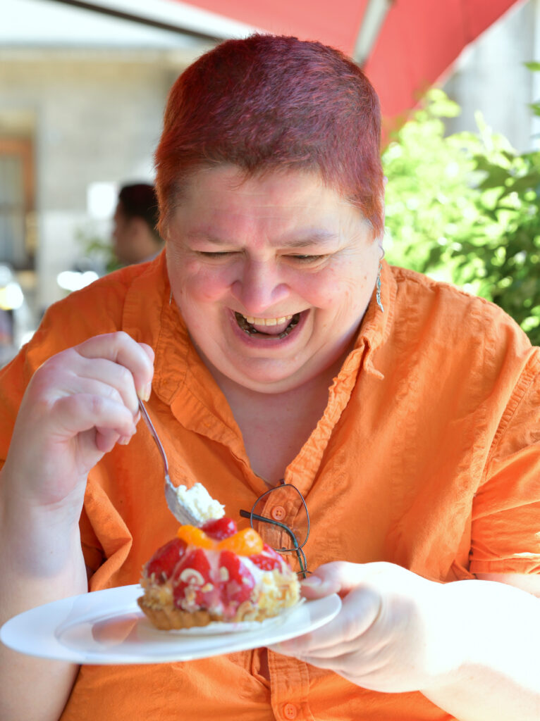 ich sitze mit lachendem Gesicht, bekleidet mit orangener Bluse im Cafe und steche gerade voller GEnuß in ein Erdbeertörtchen