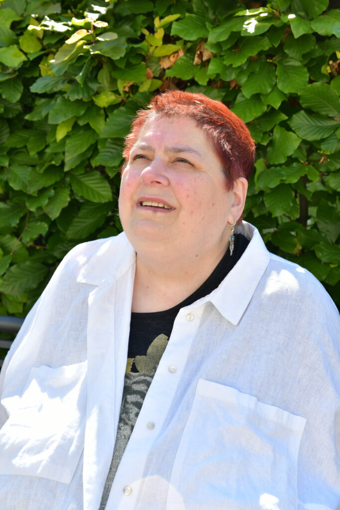 ich sitze mit einer weißen Bluse bekleidet vor einer Hecke und schaue nach oben