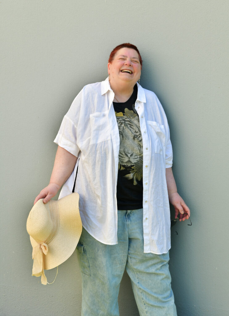 Ich stehe vor einer Wand. Ich trage eine weiße Bluse und Jeans, in der Hand einen Sonnenhut und lache gerade herzhaft