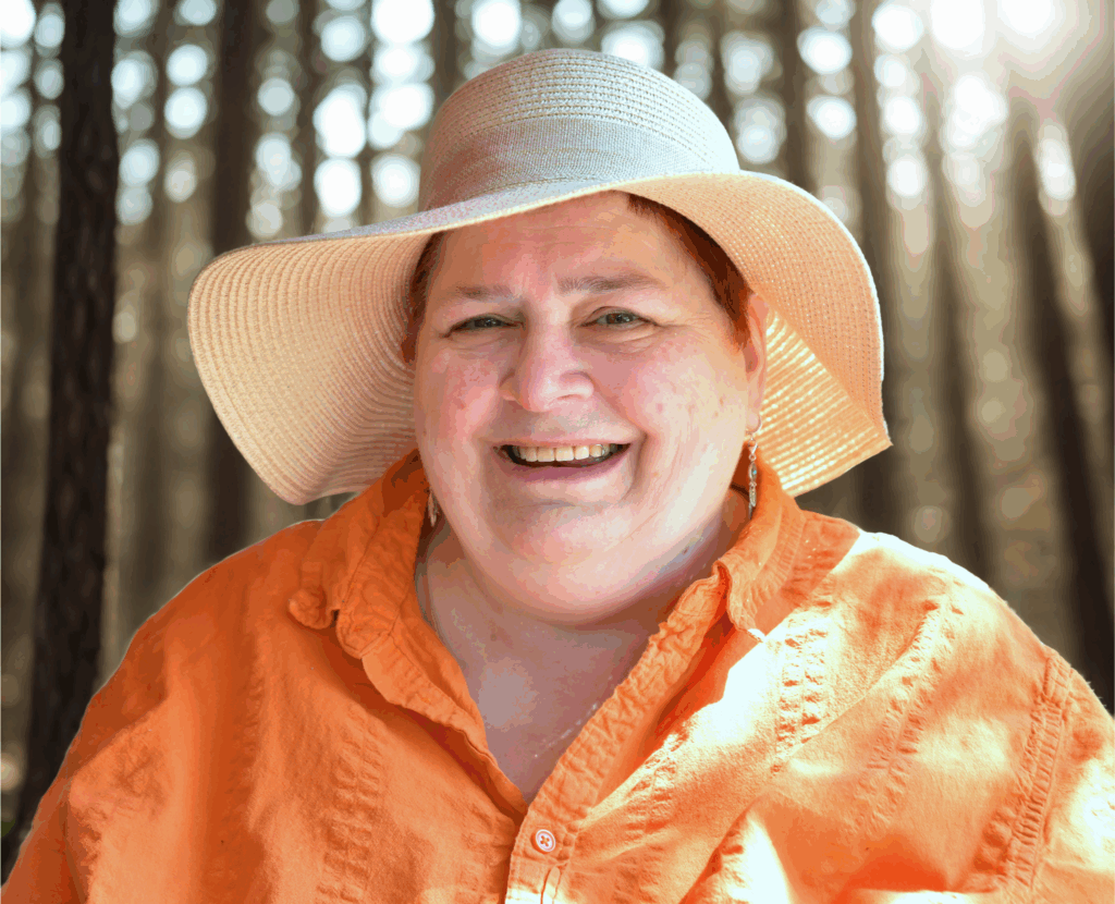Bild von mir mit orangener Bluse und großem Sonnenhut, wie immer grinsend, ich bin 61