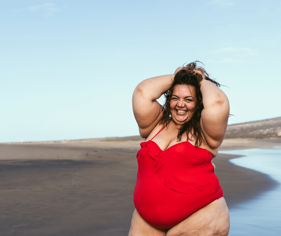Eine Frau am Strand, sie ist übergewichtig, hat aber keine Scheu sich fröhlich mit ihrem knallroten Badeanzug zu zeigen